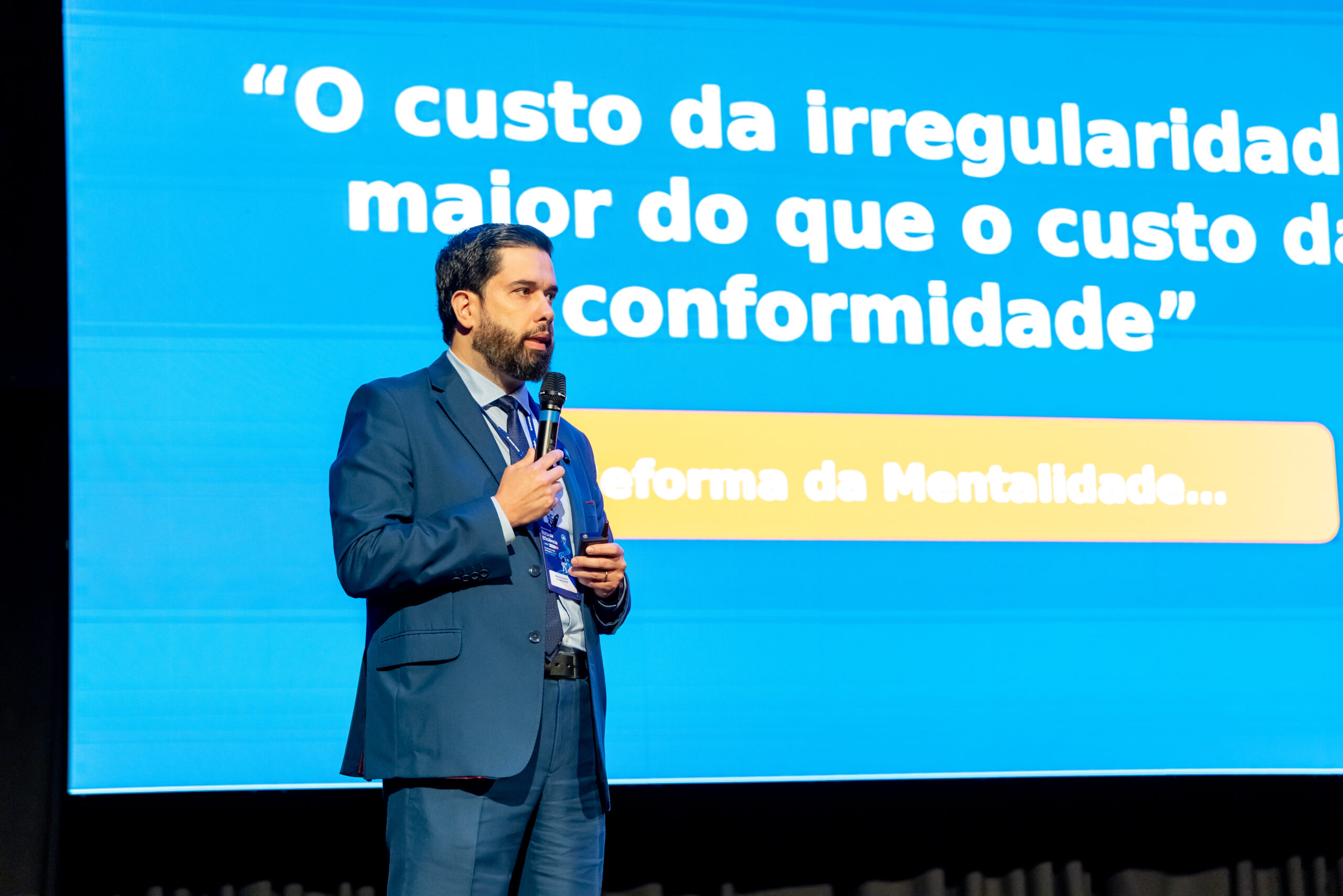Advogado tributarista Marcos Claudino palestrando em um evento, com o microfone na mão, diante de um telão azul com a frase sobre o custo da irregularidade e o custo da conformidade